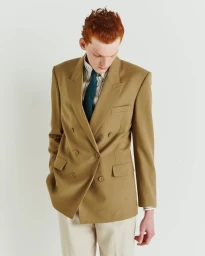 A red-haired male model wearing a camel double-breasted blazer and teal tie against a plain white studio background.