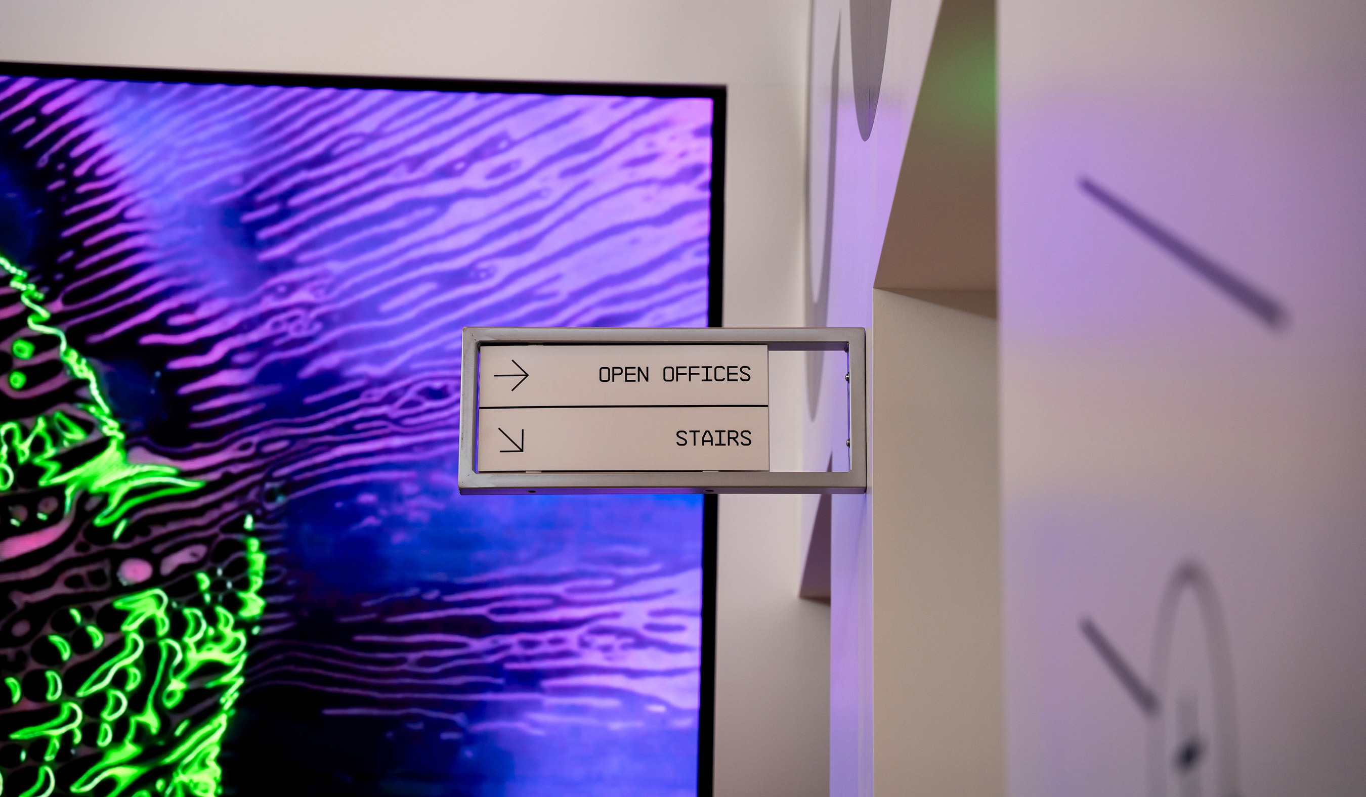 Modern directional office sign reading open offices and stairs mounted on a white wall next to a vibrant neon screen