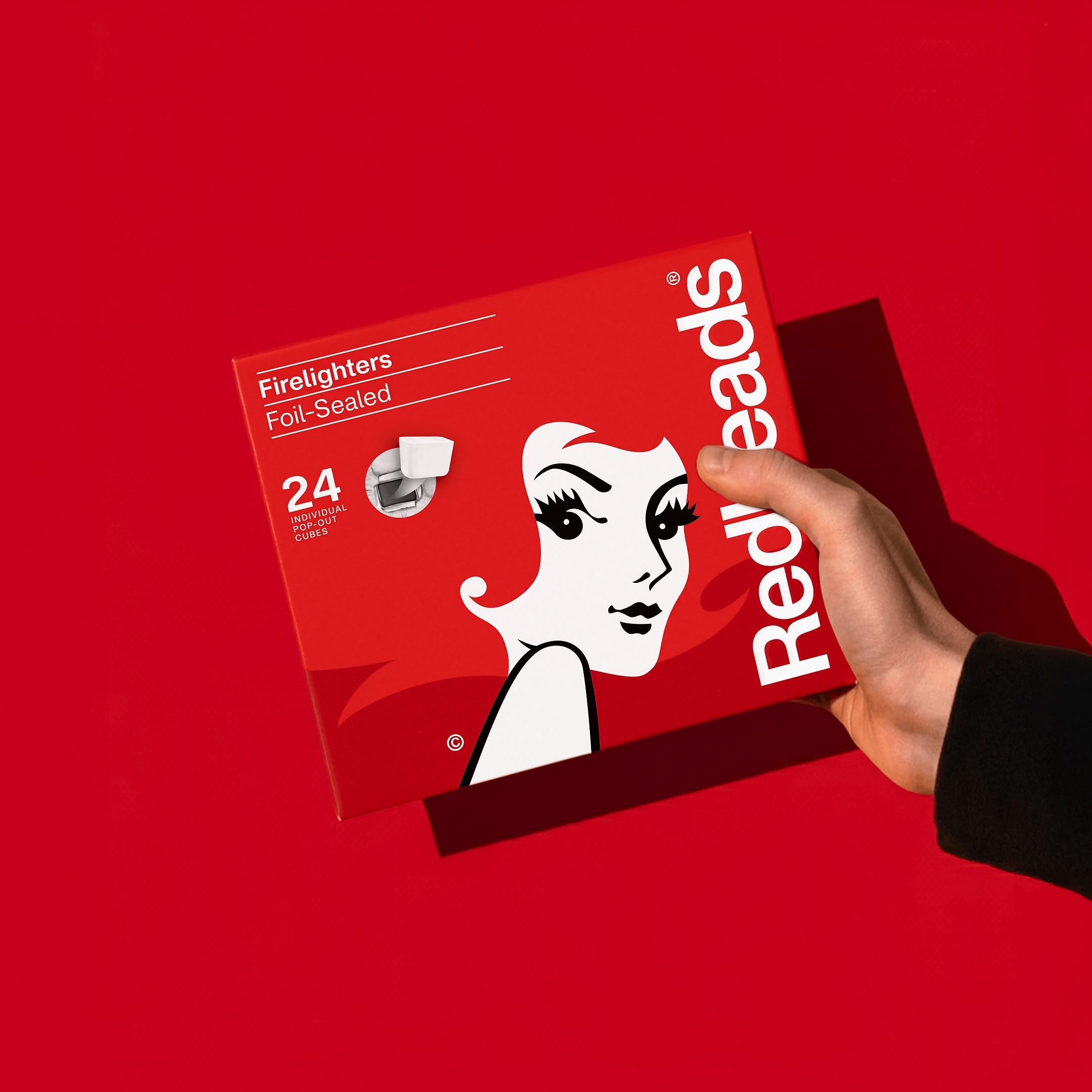 A hand holding a red Redheads firelighters box against a solid red background with a distinct shadow.