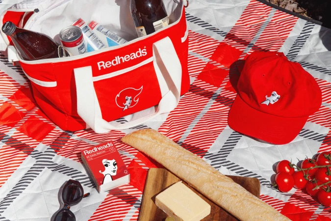 Red cooler bag, red baseball cap, matchbox, and picnic food spread out on a red and white checkered blanket.