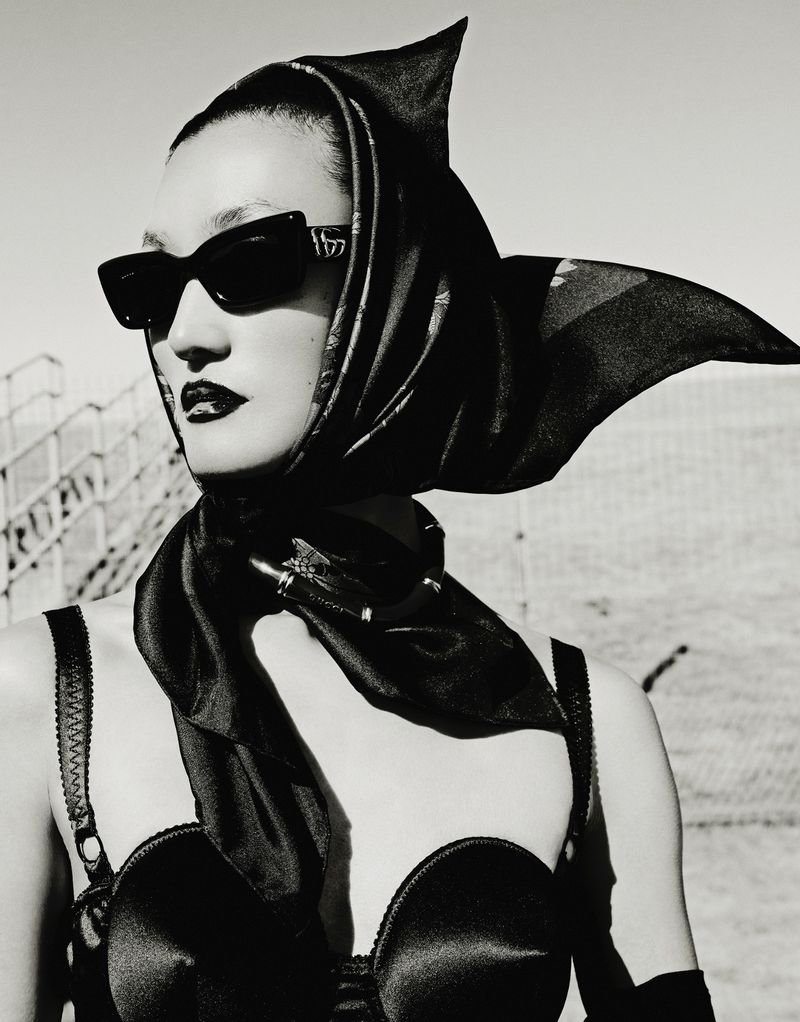 Black-and-white portrait of an adult woman with dark lipstick wearing a headscarf, sunglasses, and a bra top against a bright background.