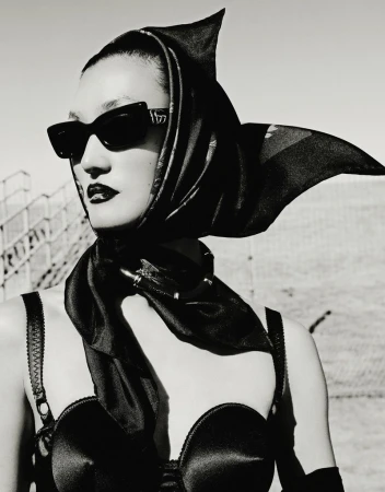 Black-and-white portrait of an adult woman with dark lipstick wearing a headscarf, sunglasses, and a bra top against a bright background.