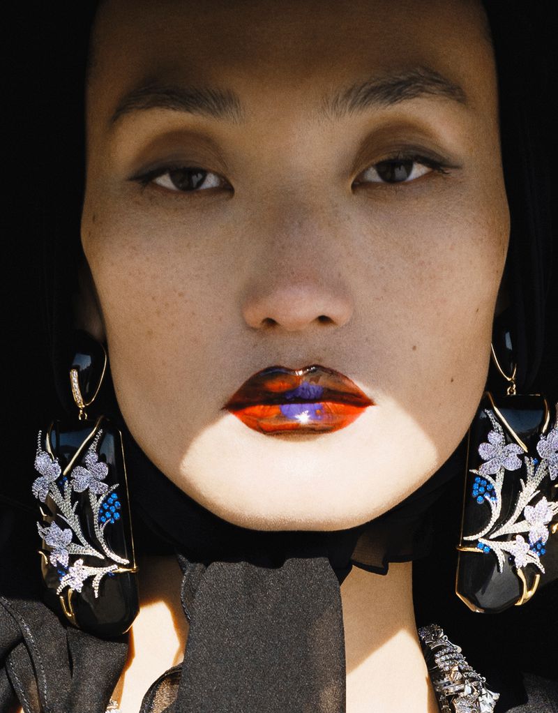 Close-up of an Asian woman's face with dramatic glossy red and purple lips and large floral earrings against a dark background.