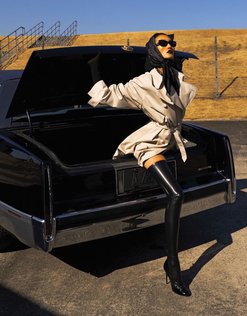 Stylish adult woman in a trench coat and thigh-high boots poses with a black vintage car on an outdoor set with neutral tones.