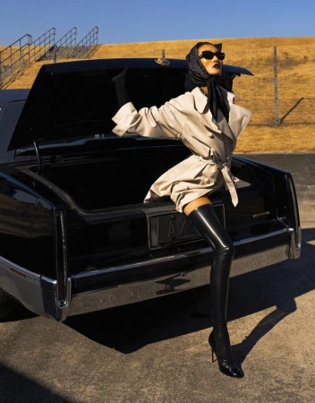 Stylish adult woman in a trench coat and thigh-high boots poses with a black vintage car on an outdoor set with neutral tones.