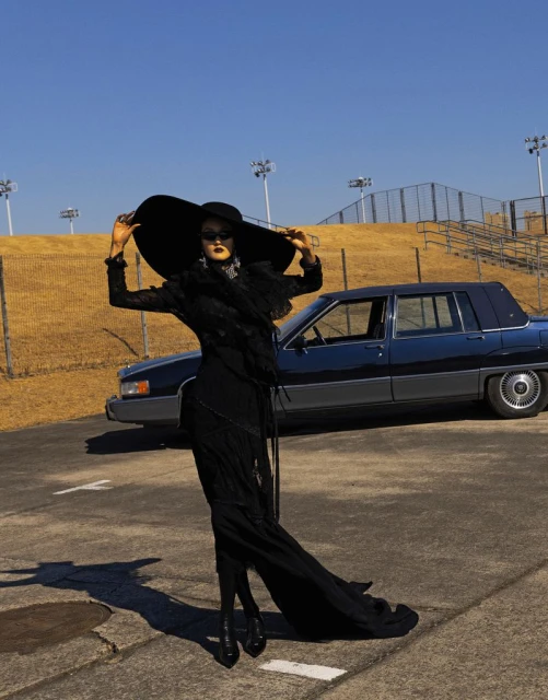 Elegant woman in black dress and wide-brimmed hat poses next to vintage car in outdoor setting under clear blue sky.