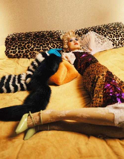 Woman in patterned dress reclines on leopard-print cushion with fur stole, patterned hose, and pale green heels indoors.