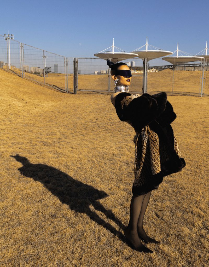 Fashionable mannequin in black and animal-print dress, black hosiery, and heels stands in a grassy field against a blue sky backdrop.