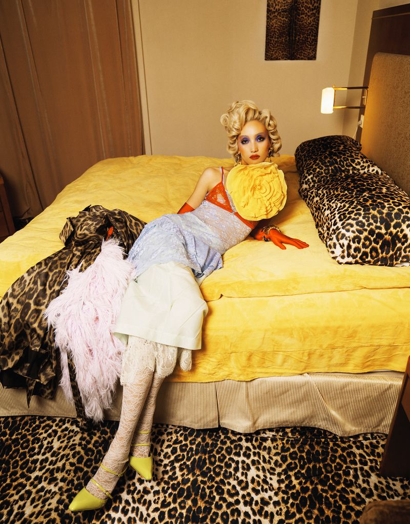Fashion photo of a woman with blonde hair, in a colorful dress and gloves, lying on a yellow bed in a leopard-print room, looking at the camera.