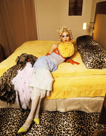 Fashion photo of a woman with blonde hair, in a colorful dress and gloves, lying on a yellow bed in a leopard-print room, looking at the camera.