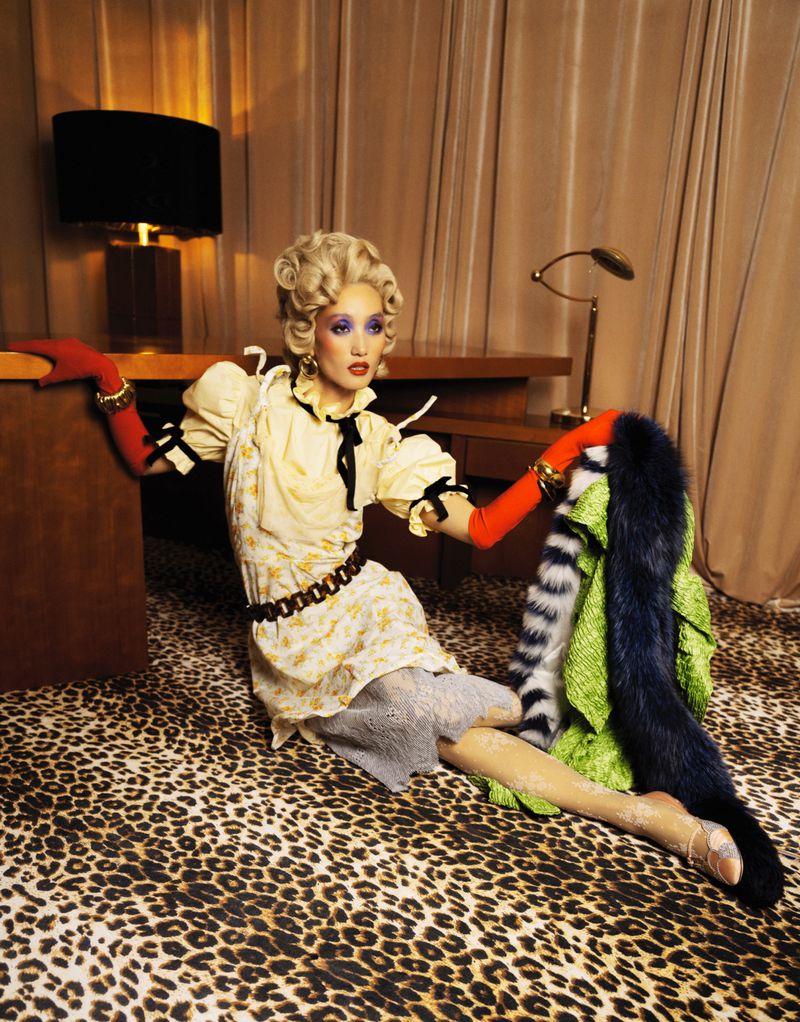 A blonde model sits on a leopard-print carpet wearing a yellow dress with red gloves near an office desk in a staged studio environment.
