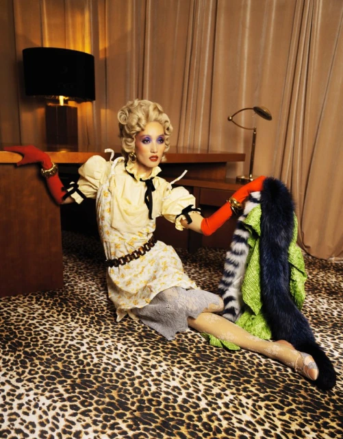 A blonde model sits on a leopard-print carpet wearing a yellow dress with red gloves near an office desk in a staged studio environment.