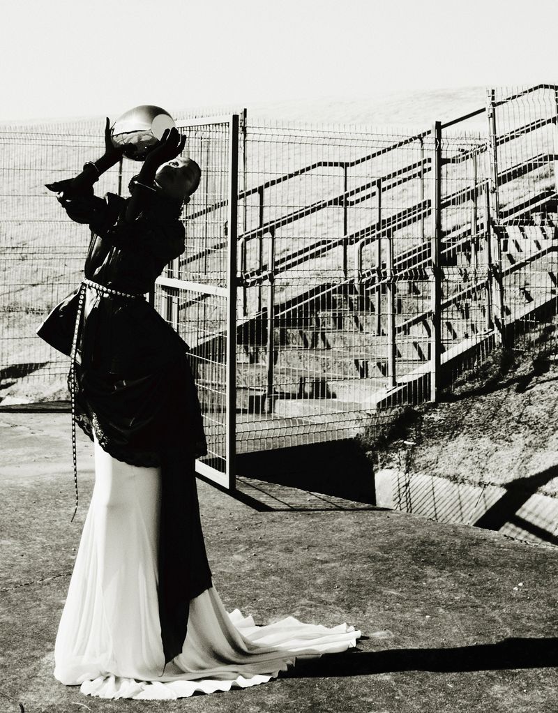 Fashion photography of a person in a black jacket and long white skirt holding a reflective ball in an outdoor urban setting.