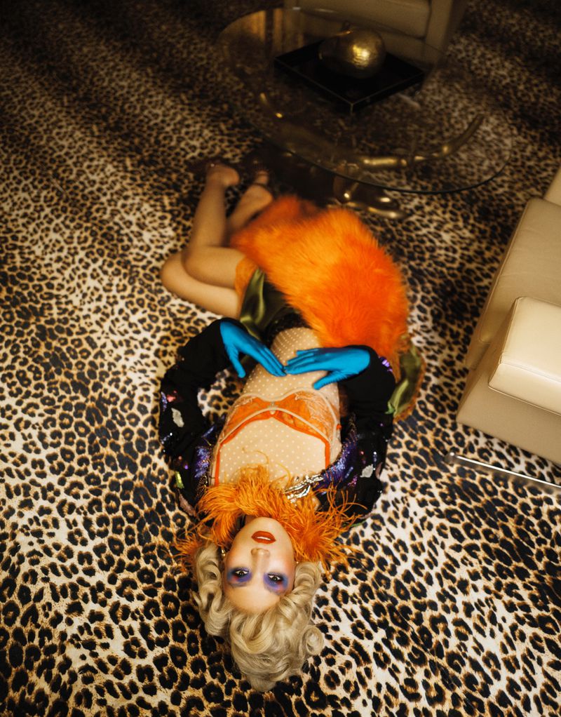 A woman with an orange feathered collar and blonde wig lies on a leopard print rug in a glamorous, retro-inspired studio setting.