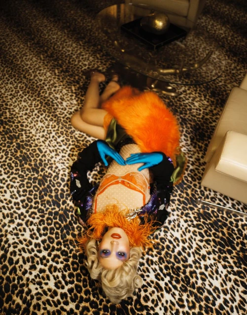A woman with an orange feathered collar and blonde wig lies on a leopard print rug in a glamorous, retro-inspired studio setting.
