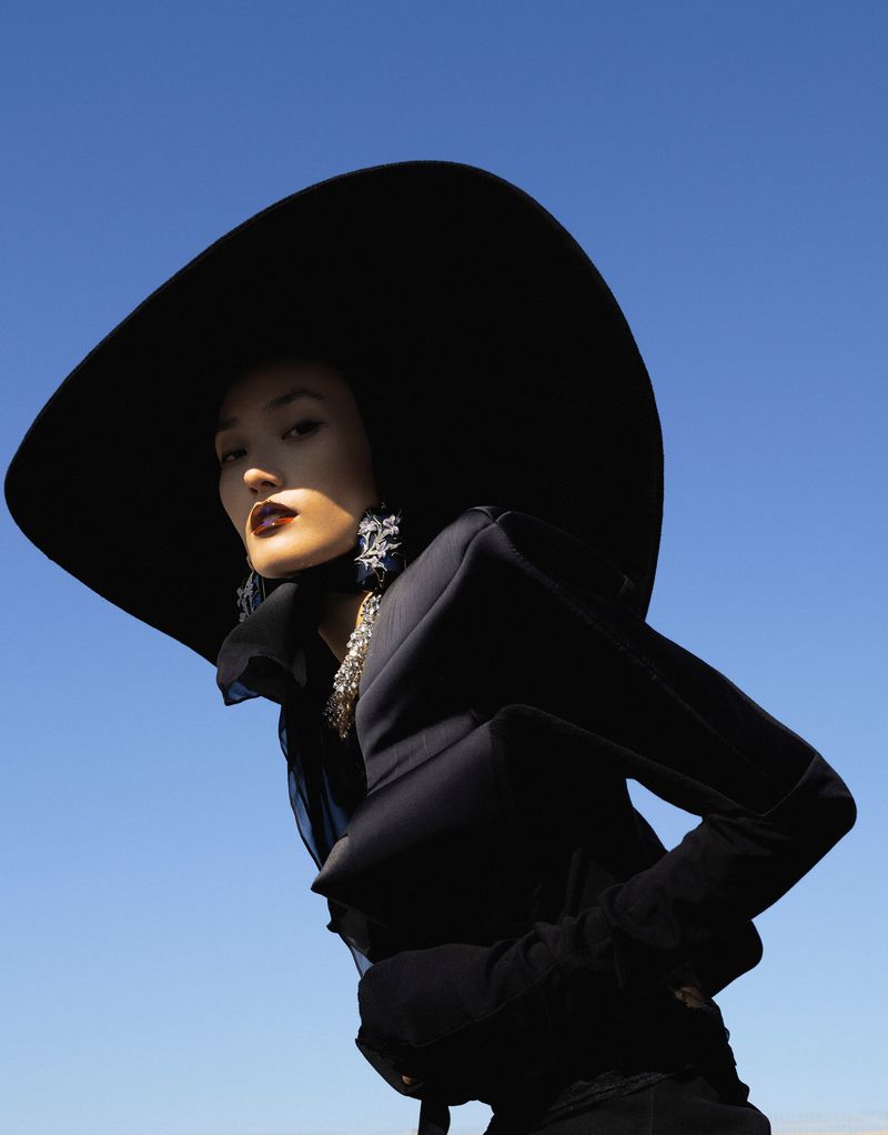 Woman poses in all-black fashion ensemble with wide-brimmed hat against a clear blue sky, low angle shot, editorial fashion.