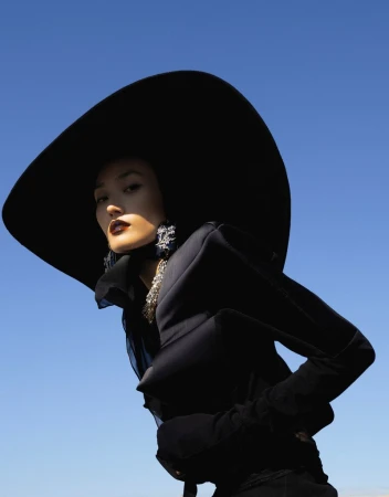 Woman poses in all-black fashion ensemble with wide-brimmed hat against a clear blue sky, low angle shot, editorial fashion.