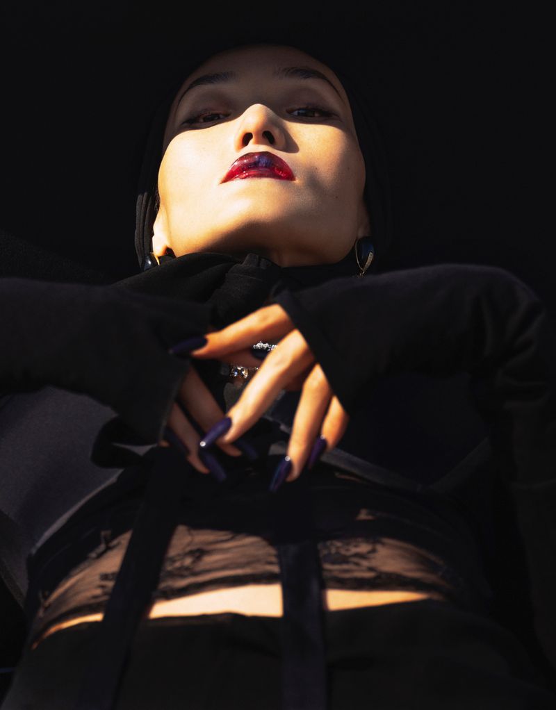 Low angle shot of a woman with dark hair and dark lipstick wearing a dark top with long sleeves against a dark background.