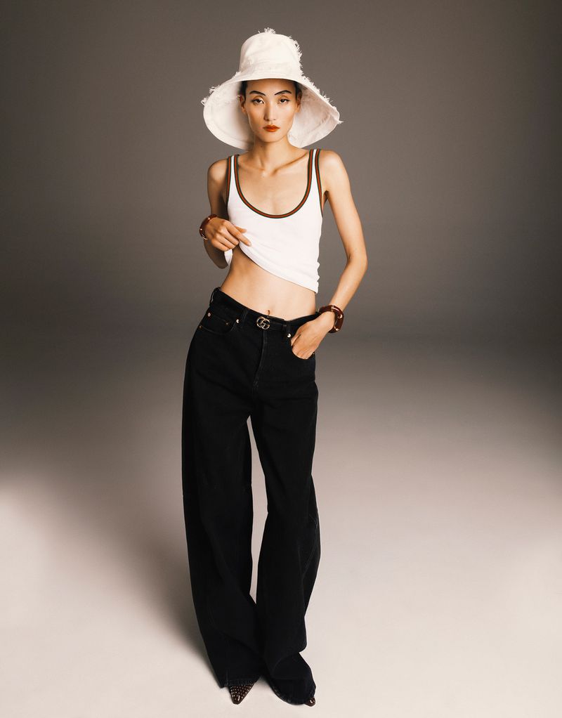 A stylish woman in a white tank top and oversized jeans poses in a studio with a white sun hat, exuding a relaxed fashion statement.