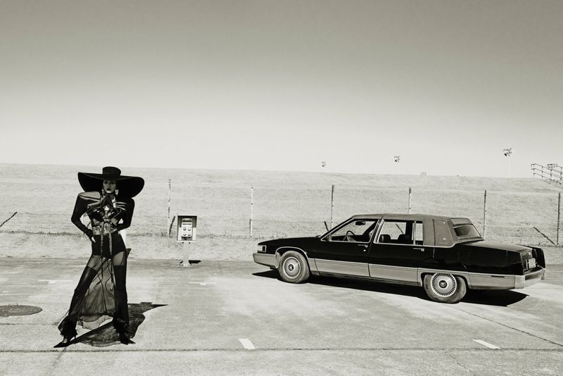 Monochrome fashion shot of a woman in avant-garde dress and large hat, standing next to an old black sedan car in an outdoor setting.