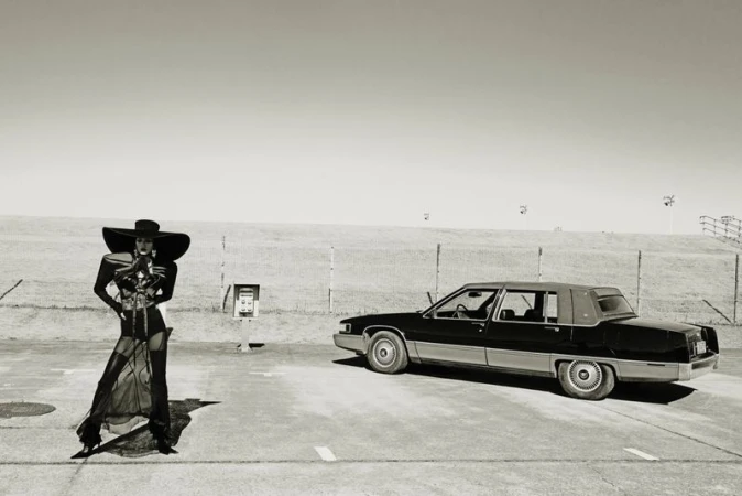 Monochrome fashion shot of a woman in avant-garde dress and large hat, standing next to an old black sedan car in an outdoor setting.