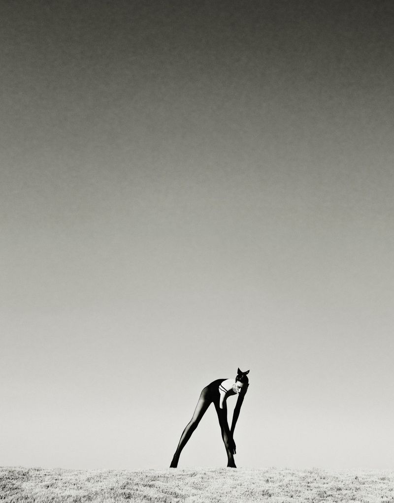Adult woman in a black one-piece swimsuit leans forward on a sand dune under a bright sky, captured in a high-contrast black-and-white shot.