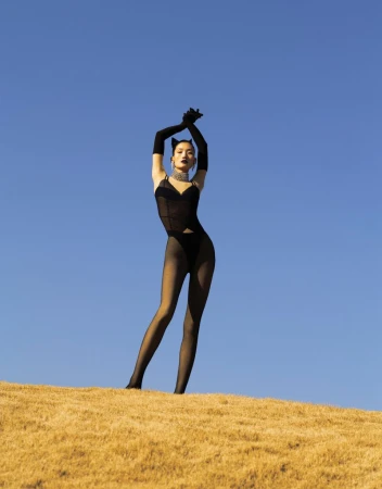 Elegant woman in a black bodysuit with long gloves poses atop a grassy hill against a bright blue sky in a fashion-forward portrait.