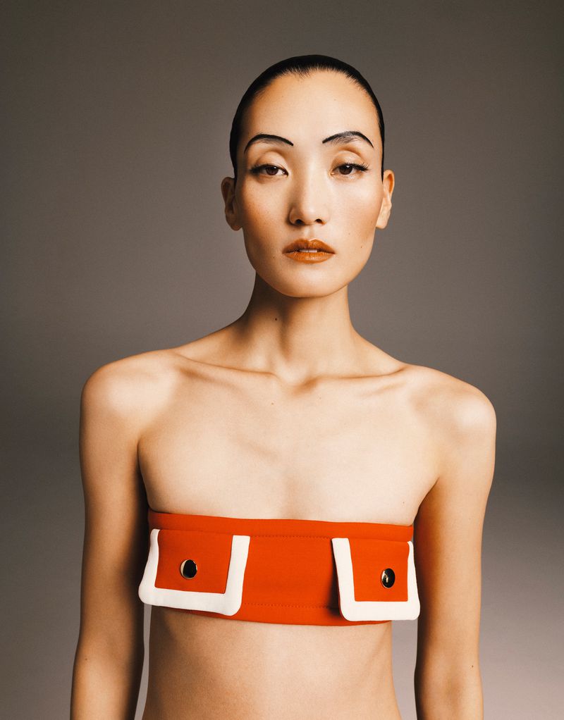 Portrait of a topless Asian woman in an orange bandeau top with white trim, featuring a makeup look with graphic drawn-on eyebrows.
