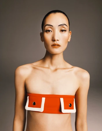 Portrait of a topless Asian woman in an orange bandeau top with white trim, featuring a makeup look with graphic drawn-on eyebrows.