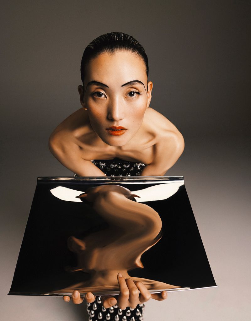 Portrait of a woman with stylized makeup and a reflective surface creating distorted reflection in a studio environment.
