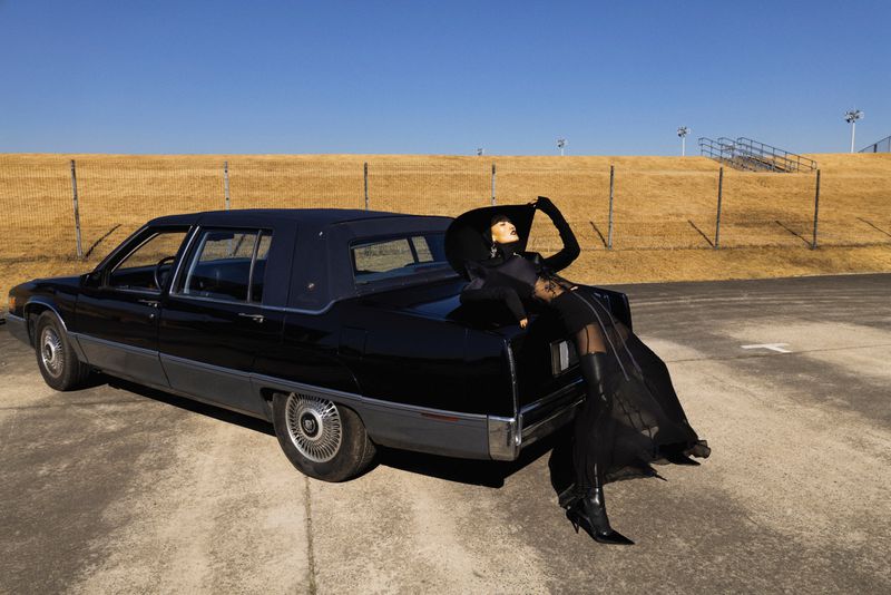 Woman in black attire and hat poses elegantly on the trunk of a vintage black car against a neutral background under a clear blue sky.
