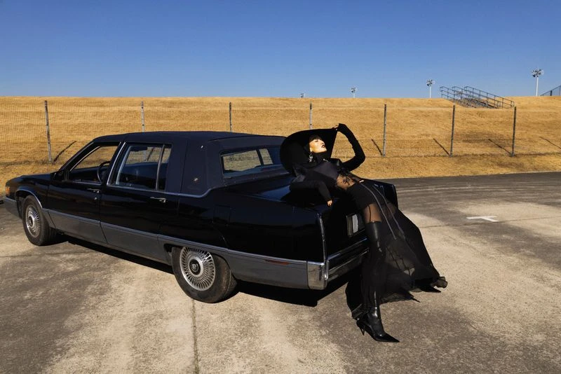Woman in black attire and hat poses elegantly on the trunk of a vintage black car against a neutral background under a clear blue sky.