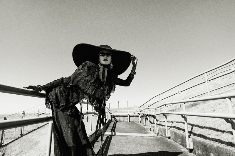 Black and white fashion shot of a model wearing a large hat and ruffled top, posing on a ramp with railings under a clear sky.