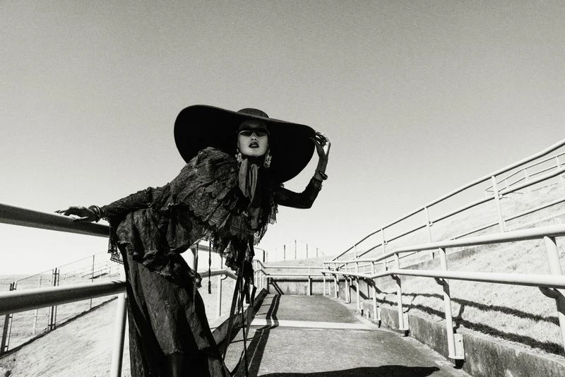 Black and white fashion shot of a model wearing a large hat and ruffled top, posing on a ramp with railings under a clear sky.