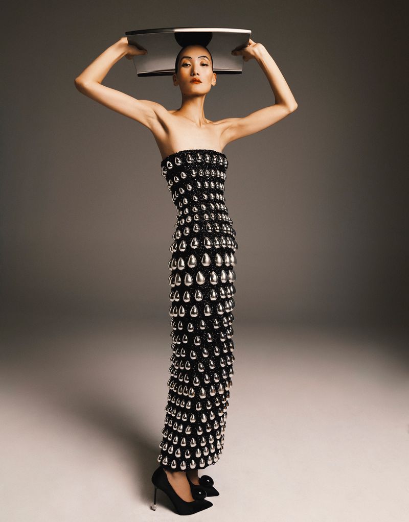 Elegant Asian woman in a black gown adorned with silver teardrop beads, balances a shiny silver tray on her head in a studio setting.