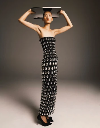 Elegant Asian woman in a black gown adorned with silver teardrop beads, balances a shiny silver tray on her head in a studio setting.