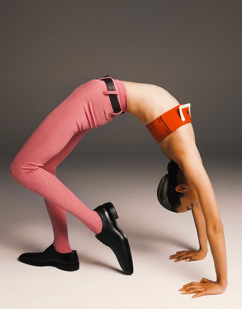 Woman in a bridge pose wearing a red belt and pink leggings with black leather shoes against a grey backdrop in a studio.