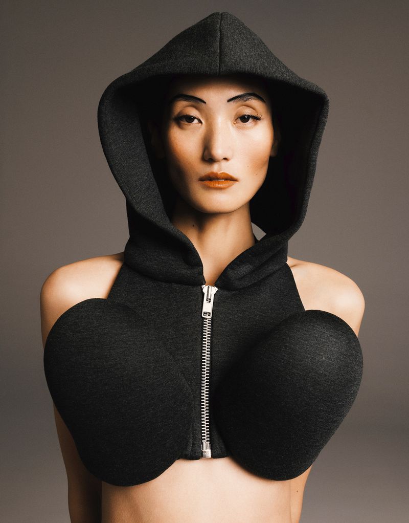 Portrait of an East Asian woman wearing a dark grey, hooded, crop top with unconventional shaping in a studio with a grey background.