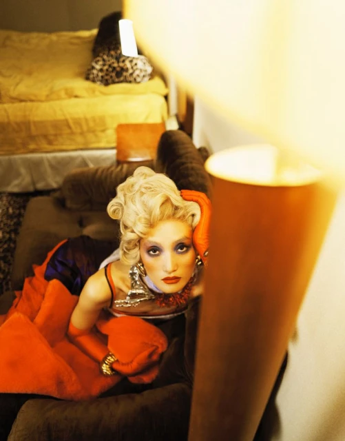Blonde woman in dramatic makeup and retro-style gown poses indoors with a serious expression and orange gloves, lit by a lamp.