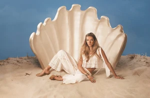 Model seated on sand in a white eyelet two-piece outfit before a giant shell prop against a blue sky