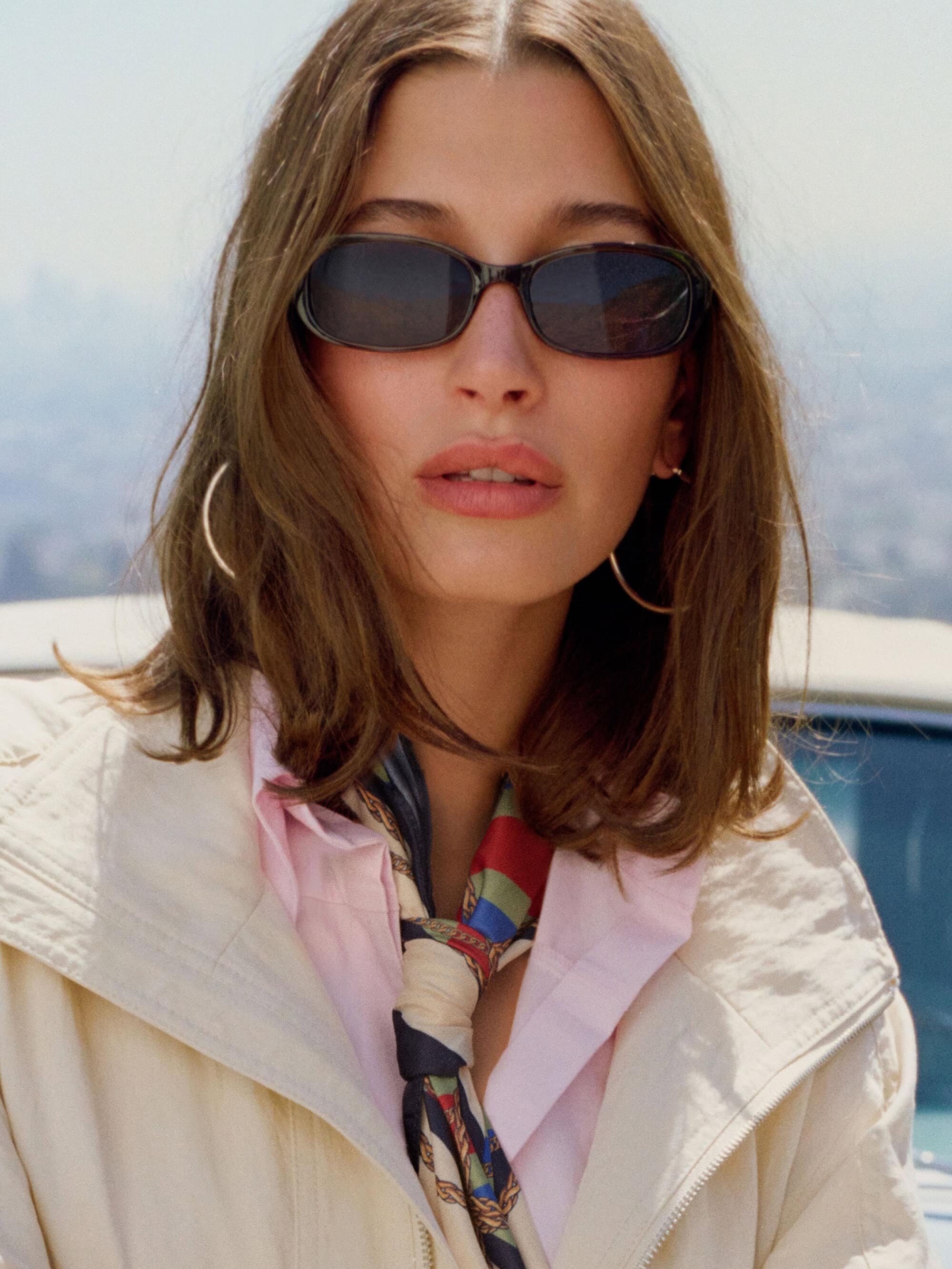 Hailey Bieber wearing sunglasses and a patterned neck scarf in a campaign portrait for Mango Summer 2026