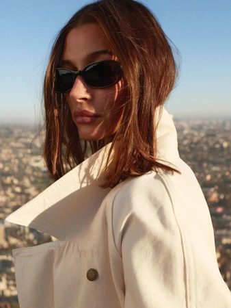 Hailey Bieber wearing sunglasses and a cream trench coat against an expansive city skyline backdrop