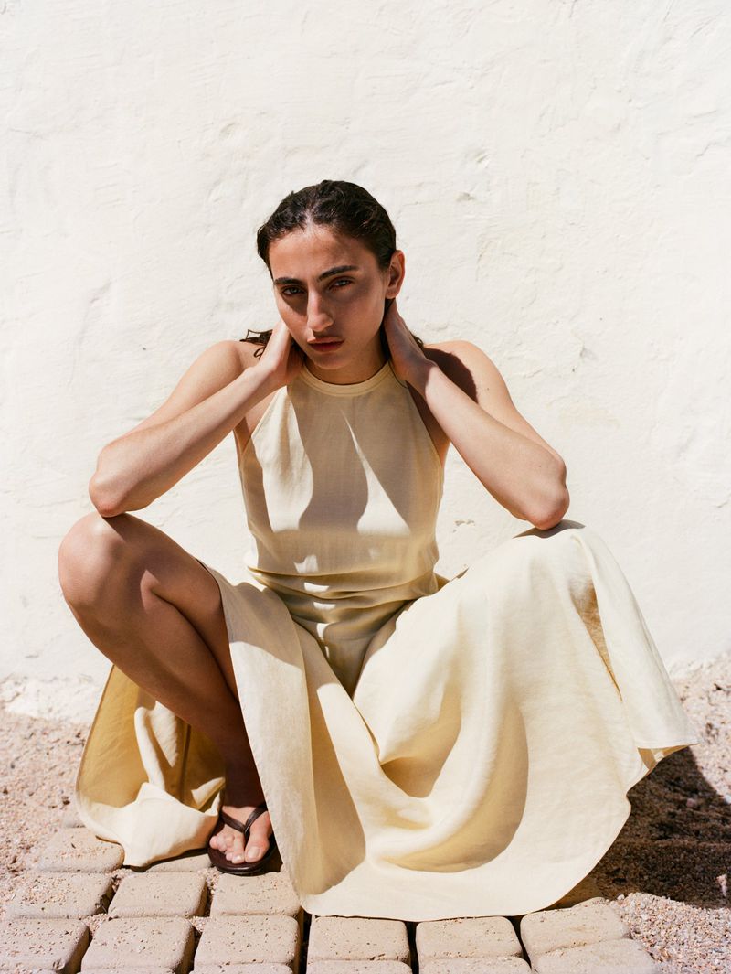 Model in a pale yellow halter dress squatting against a textured white wall for the Arket SS26 campaign