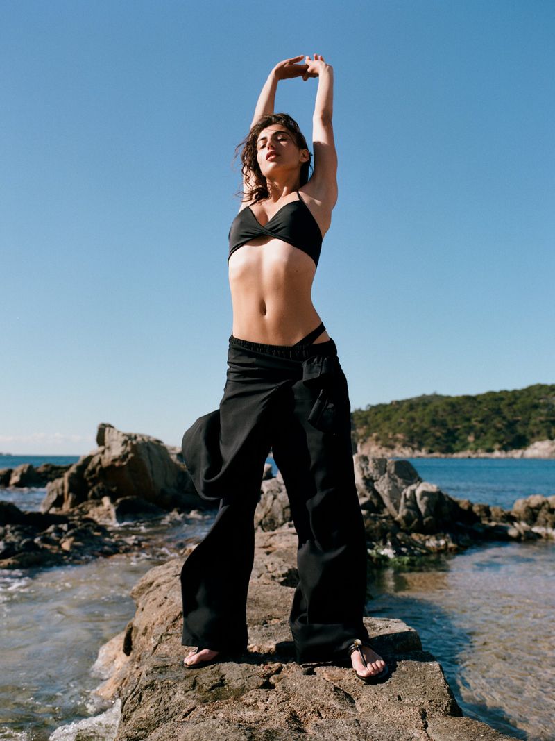 Model wearing black swimwear standing on a rocky shore with arms raised against a clear blue sky and ocean background.