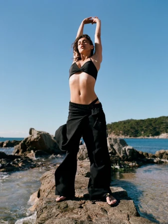 Model wearing black swimwear standing on a rocky shore with arms raised against a clear blue sky and ocean background.