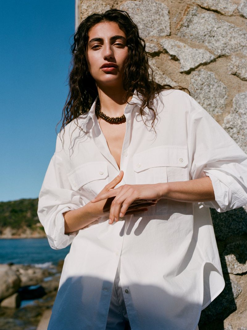 A woman with curly hair stands in a white shirt against a stone wall, with a glimpse of the ocean in the background on a sunny day.