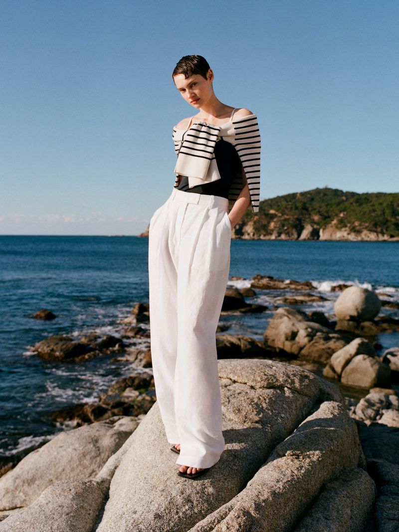 Model standing on coastal rocks wearing white trousers and a striped sweater draped over shoulders by ocean