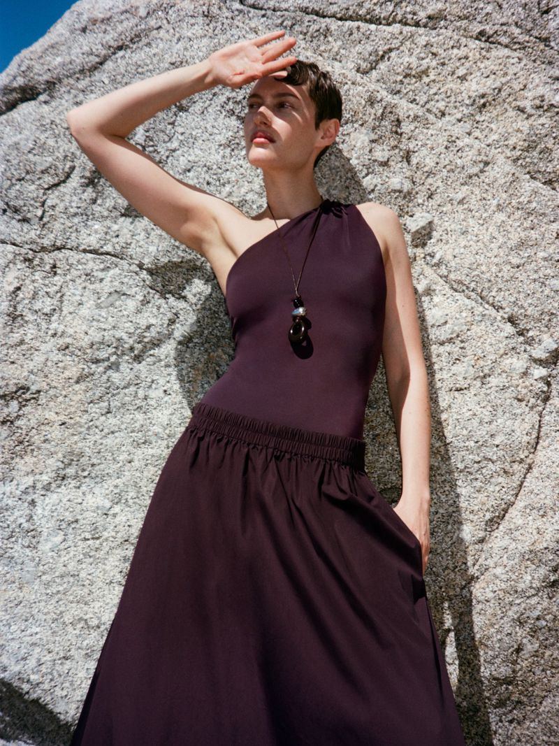 Model in a dark one-shoulder top and matching skirt shielding eyes against a rocky stone wall
