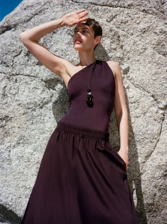 Model in a dark one-shoulder top and matching skirt shielding eyes against a rocky stone wall