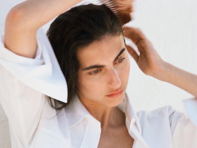 Model combing dark hair while wearing a white collared shirt against a white wall